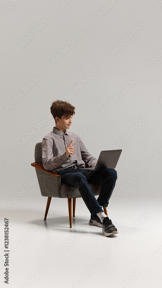 Teen boy in casual clothes sitting in armchair with laptop and having online video call, lesson against grey studio background. Concept of education, youth culture, communication, learning