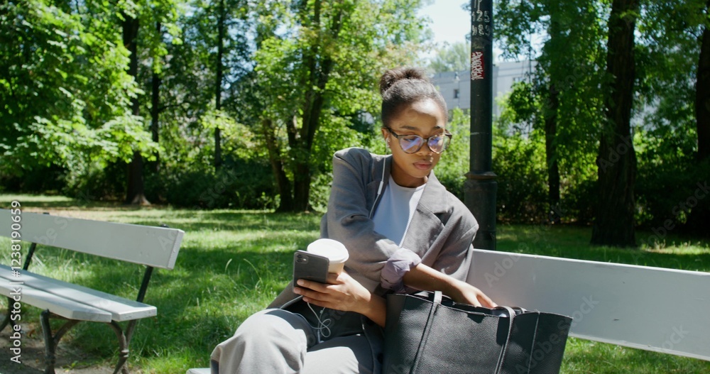 A woman in formal clothes puts on earphones and leaves a bench in a city park. She holds a mobile phone and a disposable cup of coffee in her hands A woman in formal clothes puts on earphones and leaves a bench in a city park. She holds a mobile phone and a disposable cup of coffee in her hands