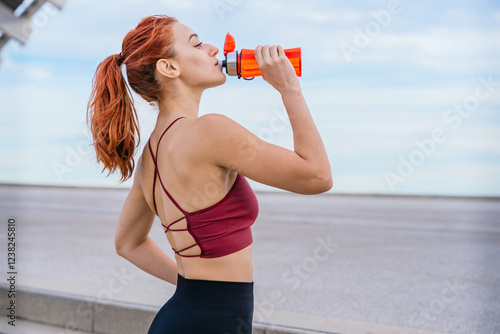 Athletic red-haired woman wearing a stylish sports bra and leggings, drinking water after exercising. She stands outdoors in an urban environment under a bright blue sky, staying hydrated and