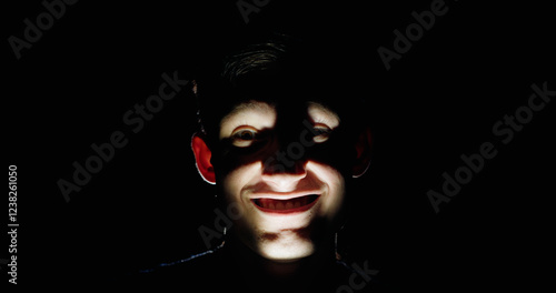 A man in darkness with an unnerving, wide smile, illuminated from below by dramatic lighting, creating eerie shadows on his face. A haunting concept of fear and suspense. photo