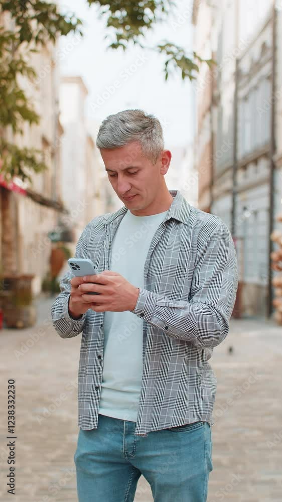 Happy smiling gray-haired man using smartphone typing text messages browsing internet social media web app working chatting online outdoor. Mature guy tourist standing in urban city street. Vertical.