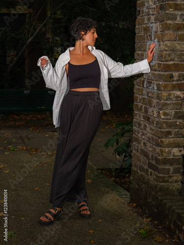 A young woman leans casually against a brick wall at night, wearing a white shirt, black crop top, wide-leg pants, and sandals, in moody lighting