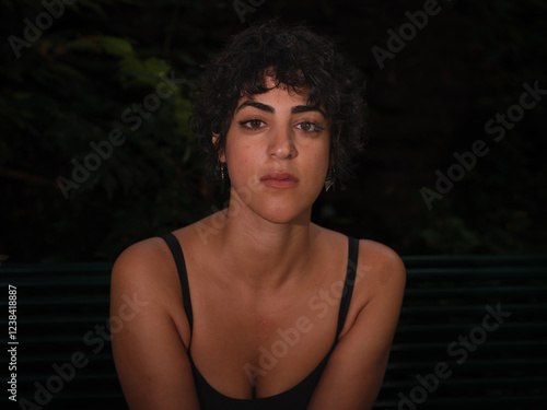 A young woman with curly hair and a serious expression looks directly at the camera, seated outdoors at night with a dark background