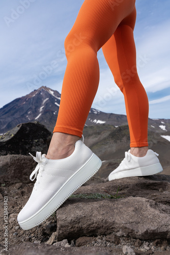 Athletic female legs wearing orange fit leggings and white sneakers. Close-up of lower part of muscular female body during outdoor fitness training in mountains with volcano in background photo