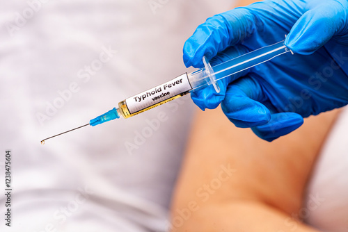 Photos The picture shows a doctor holding a syringe with the typhoid fever vaccine in her hand, wearing blue gloves, and in the background is an old woman waiting to be vaccinated with this preparation