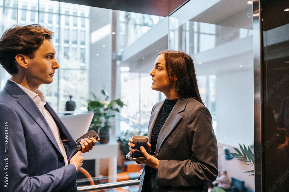 Businesswoman holding coffee cup and discussing with male colleague while standing in elevator Businesswoman holding coffee cup and discussing with male colleague while standing in elevator