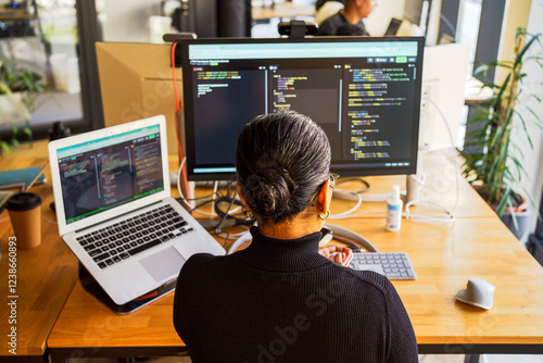 High angle rear view of female programmer coding on desktop PC and laptop while sitting at desk in office