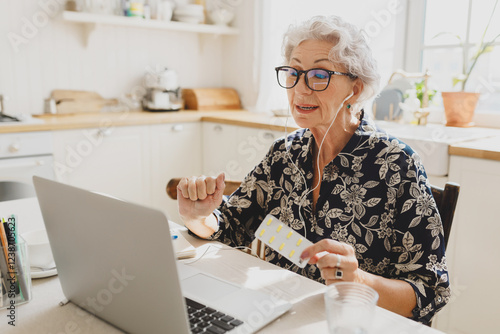 Senior pretty grandmother with gray hair in glasses consulting with her doctor online using earphones, laptop and video chat application, holding blister with pills. Telemedicine concept