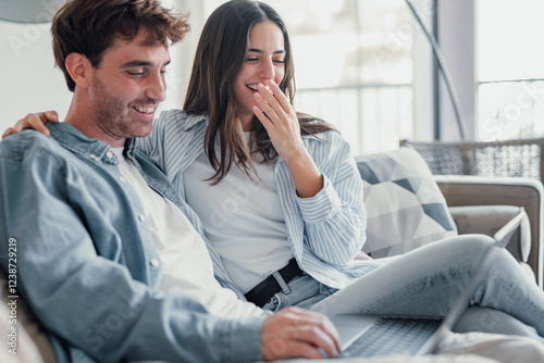 Happy millennial couple sit relax on couch in living room watching video on laptop together, smiling young husband and wife rest on sofa at home browsing Internet using modern computer device.