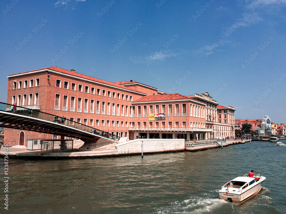 Obraz premium A View of Santa Lucia Station in Venice. The station building, with its distinctive red brick facade, is a prominent landmark along the canal. A boat passes by, adding movement and life to the scene.