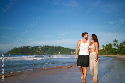 Romantic couple walks on sandy beach at sunset. Barefoot man, woman in casual summer attire hold hands, enjoying seaside honeymoon stroll. Tranquil ocean scenery, palm trees, vacation lovers leisure. photo