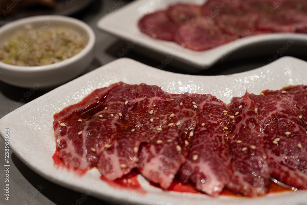 A plate of marbled raw beef slices drizzled with sauce and sprinkled with sesame seeds, ready for grilling. A side of chopped scallions is visible in the background. A plate of marbled raw beef slices drizzled with sauce and sprinkled with sesame seeds, ready for grilling. A side of chopped scallions is visible in the background.