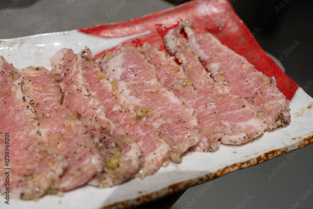 A plate of thinly sliced raw pork seasoned with black pepper, garlic, and sesame seeds, arranged on a red and white dish, ready for grilling. A plate of thinly sliced raw pork seasoned with black pepper, garlic, and sesame seeds, arranged on a red and white dish, ready for grilling.