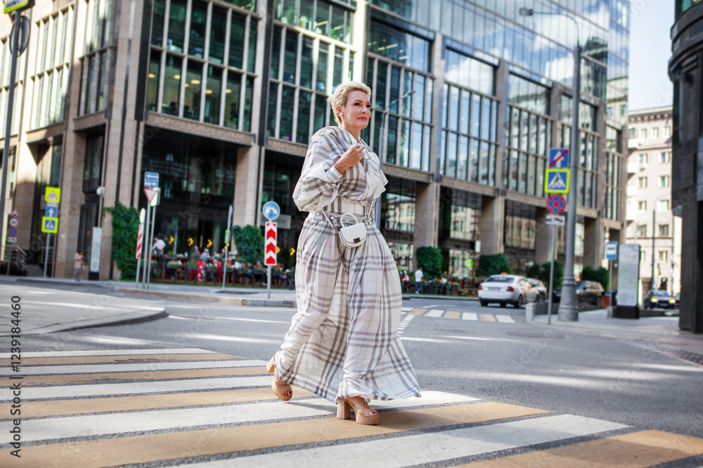 Fototapeta premium Elegant mature woman wearing white Maxi plaid dress and stylish accessories Walking confidently in urban Street near modern building, timeless fashion and sophistication. Fashion Model of Middle Age