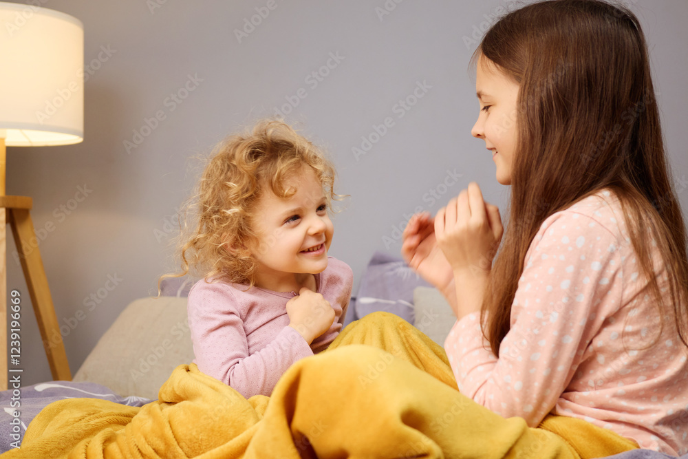 Two young sisters share laughter and joy while sitting on their bed. Warm blankets cover them as they engage in a playful conversation in a softly lit bedroom Two young sisters share laughter and joy while sitting on their bed. Warm blankets cover them as they engage in a playful conversation in a softly lit bedroom