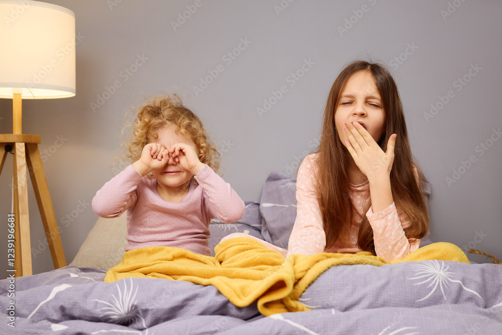 Two young girls are sitting on a bed in a cozy bedroom. One girl has curly hair and rubs her eyes, while the other yawns, both appearing sleepy and tired as they greet the morning Two young girls are sitting on a bed in a cozy bedroom. One girl has curly hair and rubs her eyes, while the other yawns, both appearing sleepy and tired as they greet the morning