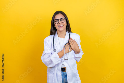 Young brunette doctor woman wearing stethoscope standing over isolated yellow background smiling with her hands on her chest and grateful gesture on her face.