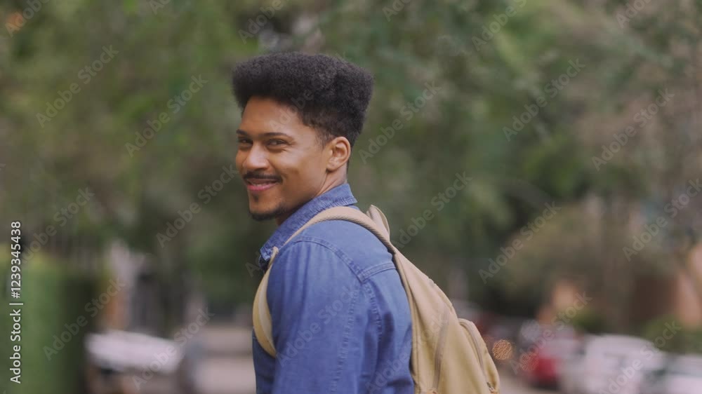 Smiling young man with a backpack turns to look back at the camera while walking outdoors, continuing on his path without stopping. Smiling young man with a backpack turns to look back at the camera while walking outdoors, continuing on his path without stopping.