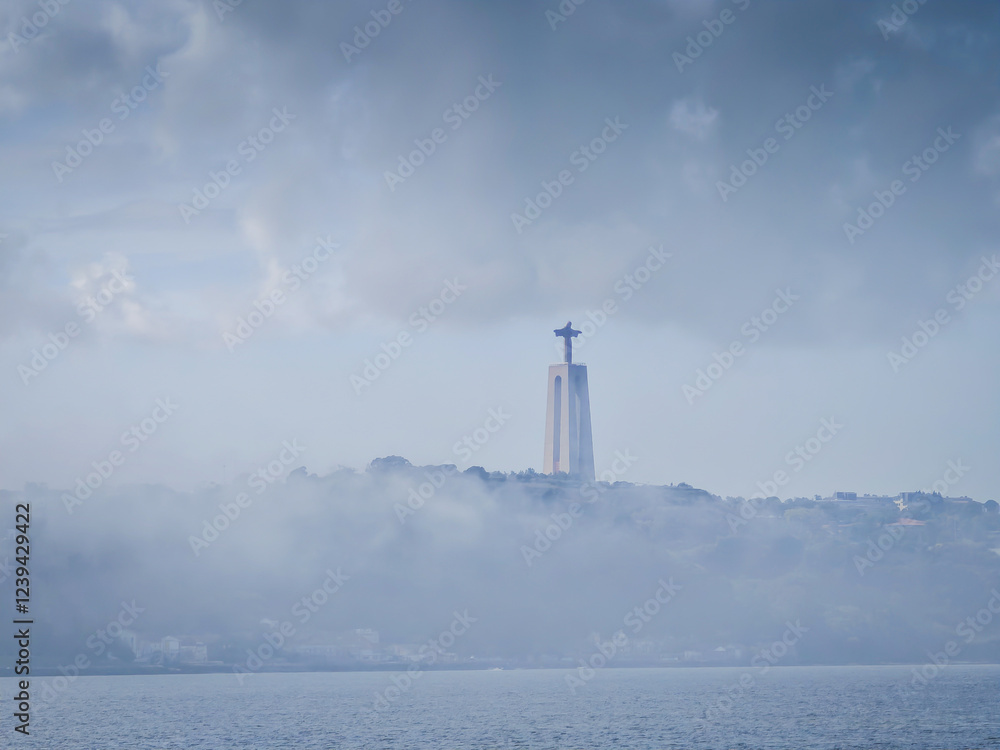Fototapeta premium Cristo Rei Statue in the fog next to 25 April Bridge near Tagus river. Sanctuary of Christ the King on a cloudy day