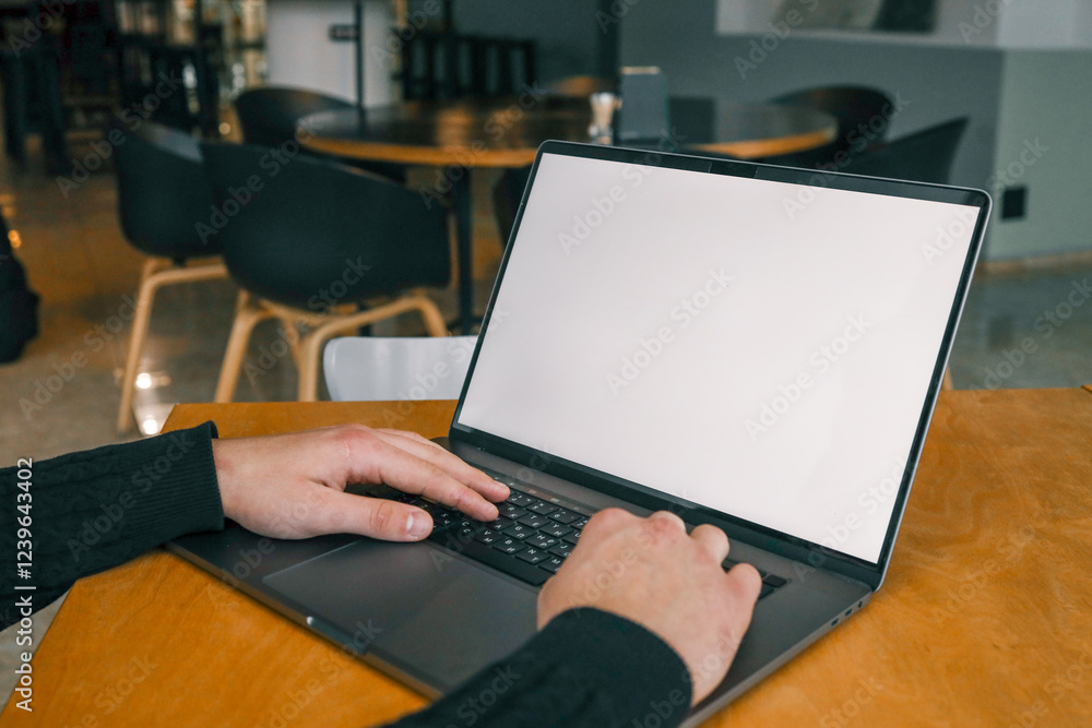 Hands typing on a laptop with a blank white screen, placed on a wooden desk in a modern cafe setting, with blurred chairs and a table in the background Hands typing on a laptop with a blank white screen, placed on a wooden desk in a modern cafe setting, with blurred chairs and a table in the background
