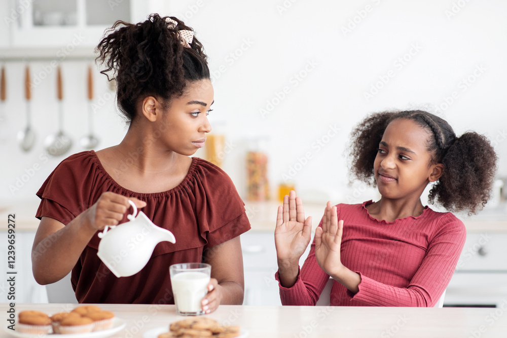 Lactose intolerance concept. Black teen girl refuse to drink milk, sitting at kitchen table with her mom, eating homemade pastry, african american woman mother pouring milk into glass Lactose intolerance concept. Black teen girl refuse to drink milk, sitting at kitchen table with her mom, eating homemade pastry, african american woman mother pouring milk into glass