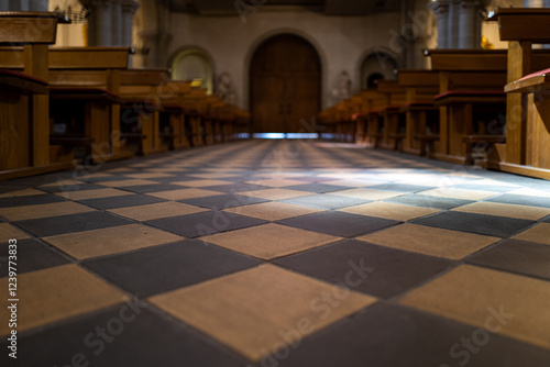 Sunlight streams onto the checkered tile floor of a church, illuminating rows of wooden pews leading towards a grand entrance. photo