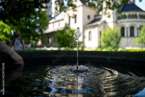 A single water droplet falls into a tranquil fountain, creating ripples in the water's surface, with an old church subtly in the background. photo