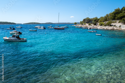 Fototapeta Naklejka Na Ścianę i Meble -  Tranquil turquoise bay with multiple boats gently anchored, surrounded by lush greenery under a clear blue sky.