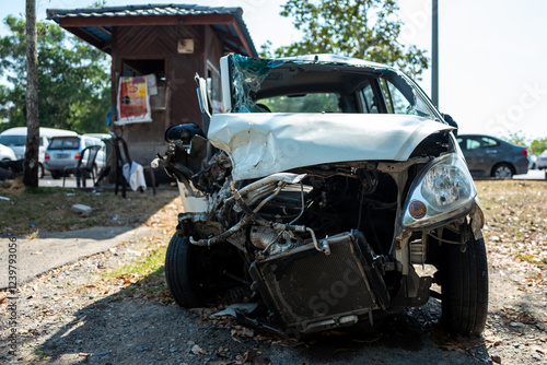 A wrecked white car sits on the roadside, its front end severely damaged in a serious accident, the windshield shattered, and the engine exposed.