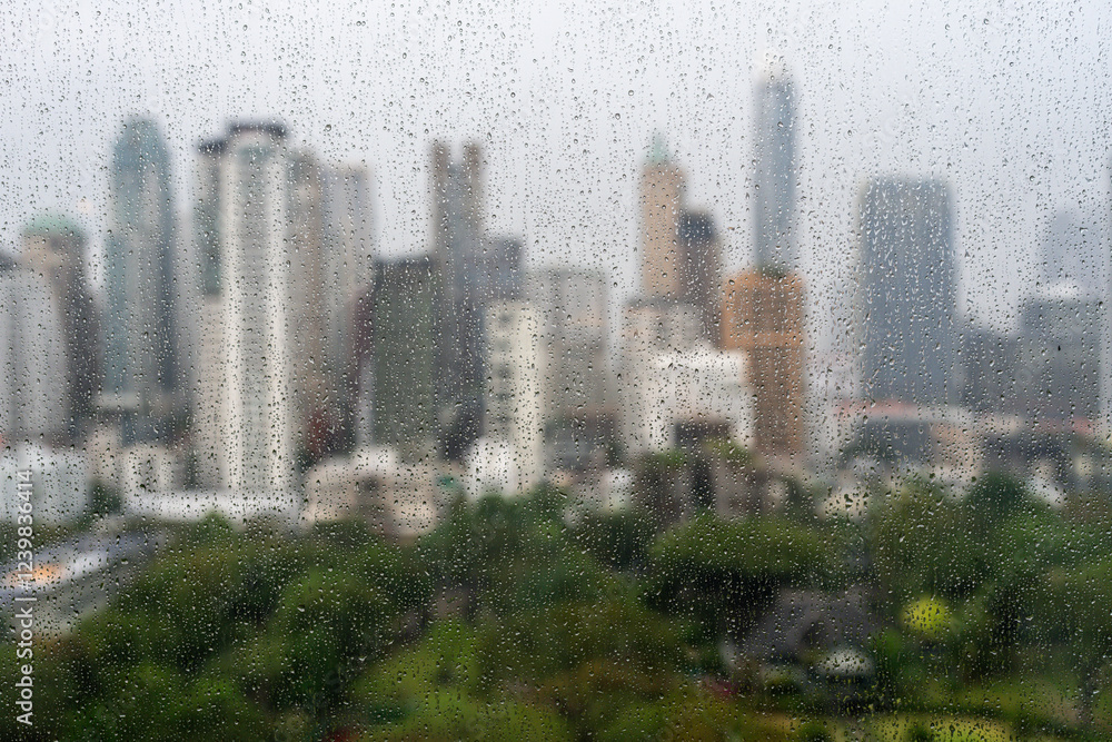 Rainy day view of a city seen through a rain-covered window, showcasing blurred skyscrapers and lush greenery.