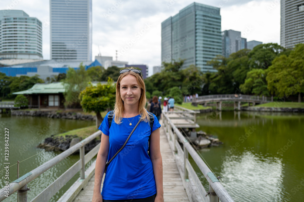 A woman in a blue dress walks on a bridge in a serene Japanese garden, modern skyscrapers rising in the background. A woman in a blue dress walks on a bridge in a serene Japanese garden, modern skyscrapers rising in the background.