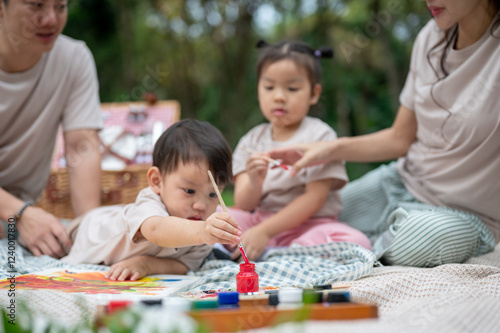 A cute little Asian boy enjoys painting on a canvas while picnicking with his parents and sister.