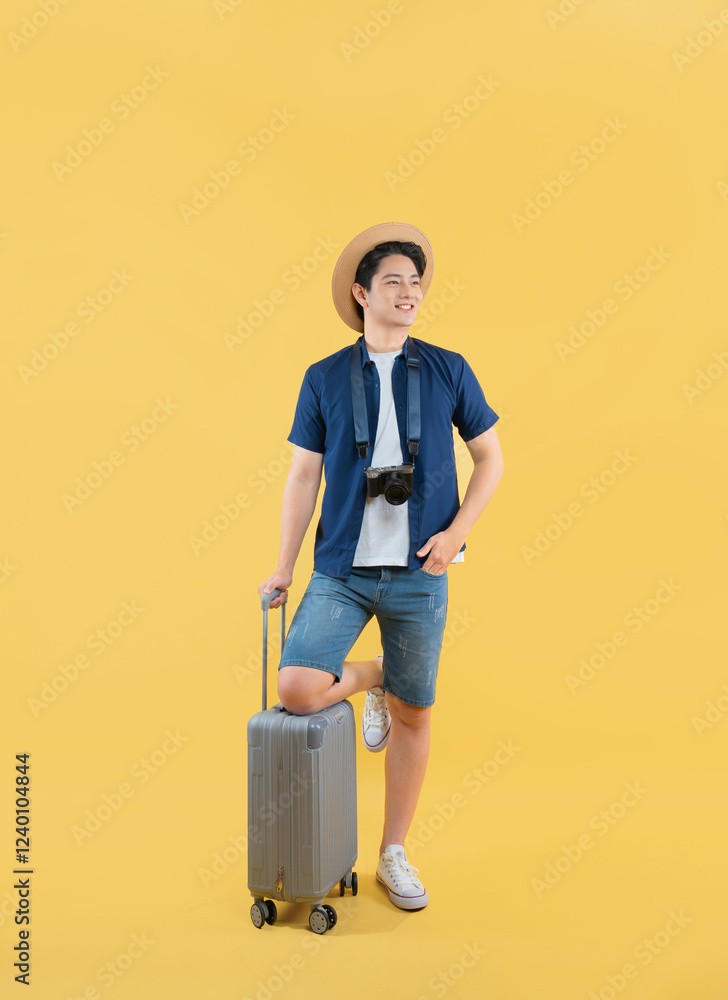 Full body image of a young Asian man pulling a suitcase, wearing a travel camera and posing on a yellow background Full body image of a young Asian man pulling a suitcase, wearing a travel camera and posing on a yellow background