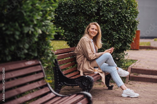 Young blonde woman with a smartphone and coffee resting on a bench in a city park