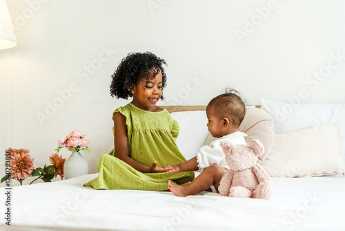 A loving moment between two African sisters sitting on a cozy bed, playing and bonding with a teddy bear. The warm, minimal bedroom setting enhances the theme of childhood, family love, and happiness. photo