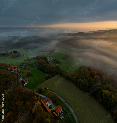 Aerial view of misty sunrise over dense forest and green valley with homes and fields, Bad Loipersdorf, Austria. photo