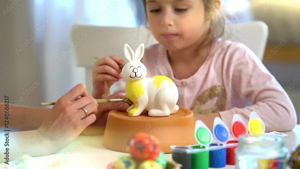 Mother and daughter paint a ceramic bunny together for Easter. The girl, wearing bunny ears, carefully applies yellow paint while her mother assists. The table is filled with colorful paints Mother and daughter paint a ceramic bunny together for Easter. The girl, wearing bunny ears, carefully applies yellow paint while her mother assists. The table is filled with colorful paints
