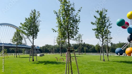 Bratislava, Bratislava Region, Slovak Republic - 09.07.2024: A sunny park with vibrant green grass, trees, and a colorful modern sculpture of large interconnected spheres.