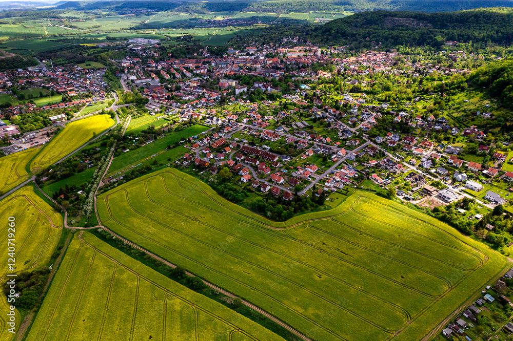 Top view of a field of yellow rapeseed flowers with a small town in the background. The sky is cloudy and the sun is shining through the clouds. Germany.