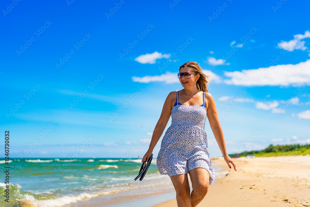 Summertime by sea. Beautiful middle aged woman tourist walking on sandy beach on sunny summer day. Front view Summertime by sea. Beautiful middle aged woman tourist walking on sandy beach on sunny summer day. Front view