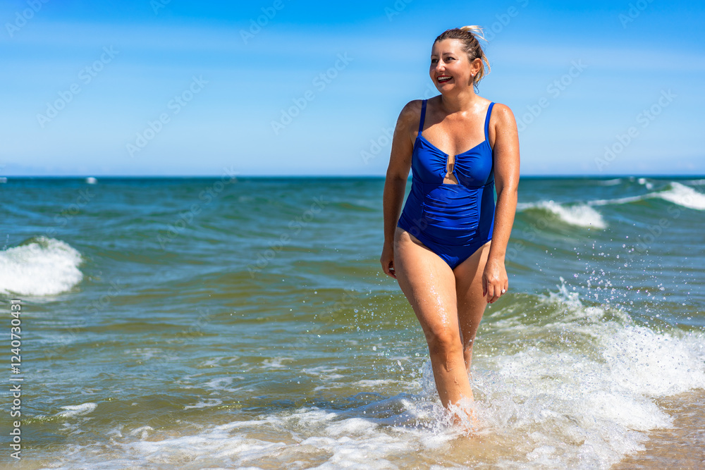 Fototapeta premium Summer vacations. Beautiful mature woman in swimsuit walking in shallow water in sea. Front view
