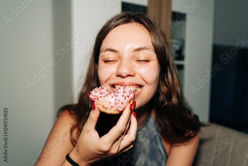 A cheerful young woman bites into a pink-frosted donut topped with sprinkles, enjoying a sweet treat indoors