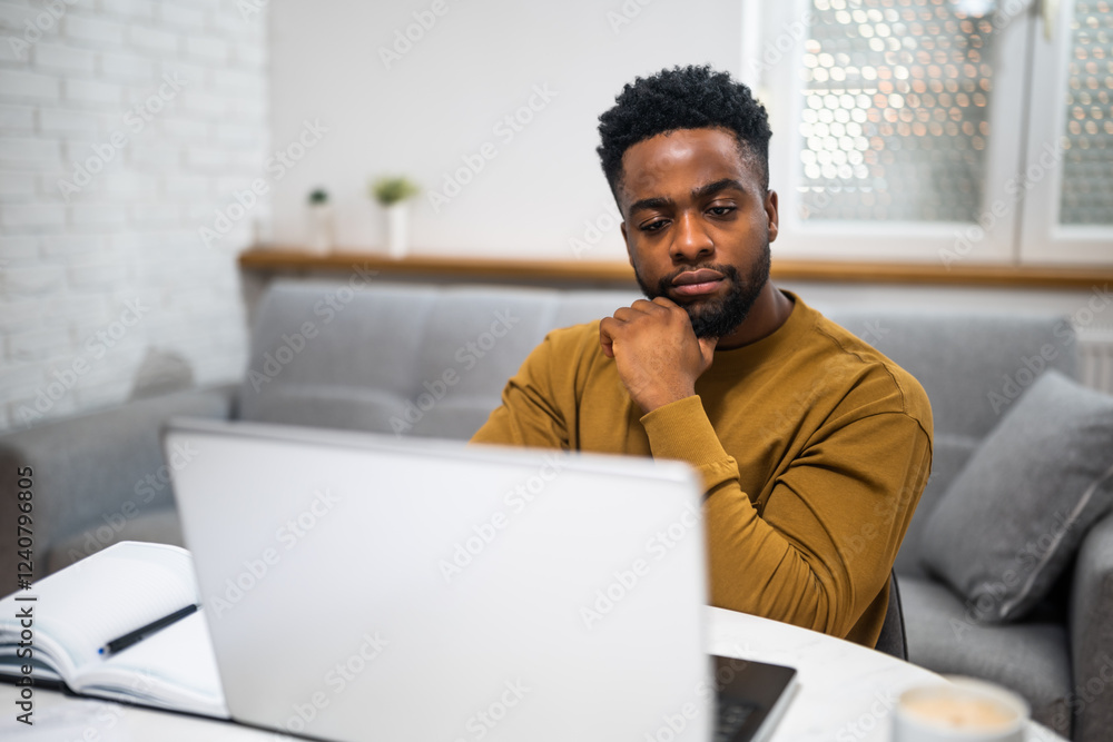 Confident and concentrated black businessman sitting at the table while using laptop and working remotely at his cozy home. Confident and concentrated black businessman sitting at the table while using laptop and working remotely at his cozy home.