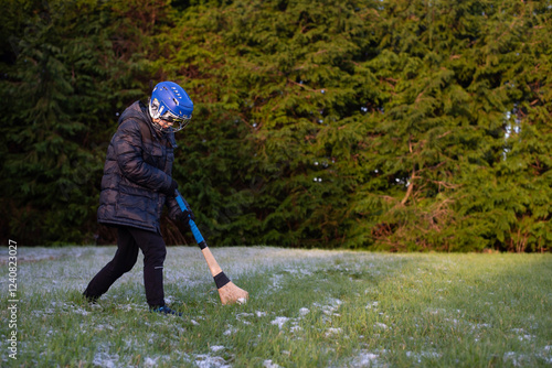 A young boy wearing a blue helmet and coat practices with hurley on frosty grass surrounded by greenery. A boy practices hurling outdoor in winter photo