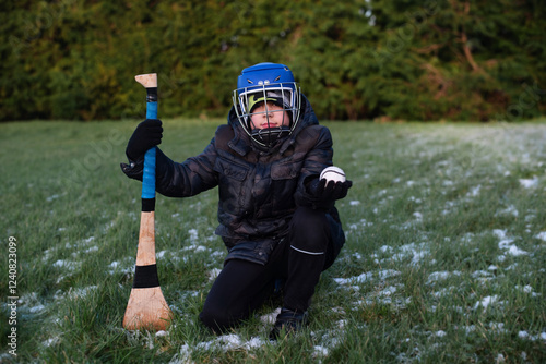 A young hurling player in protective gear holding a hurl and sliotar, kneeling on a frosty grass field during winter, signifying dedication, sportsmanship, and passion for hurling in Ireland photo