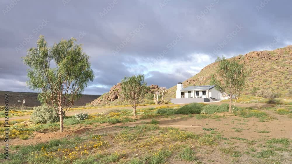 Remote guest farm in the Karoo with a white house, trees, and rocky hills under a cloudy sky Remote guest farm in the Karoo with a white house, trees, and rocky hills under a cloudy sky
