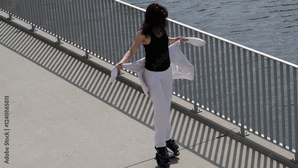 A stylish woman pauses near the water, holding her jacket while standing on rebound shoes. The combination of fitness and fashion highlights modern trends in active and mindful living. A stylish woman pauses near the water, holding her jacket while standing on rebound shoes. The combination of fitness and fashion highlights modern trends in active and mindful living.