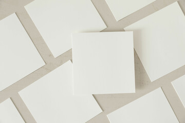 Many empty white square business card greeting card mockups with soft shadows lying diagonally on beige textured wooden table background. One card above. Flat lay, top view. Stationery composition.
