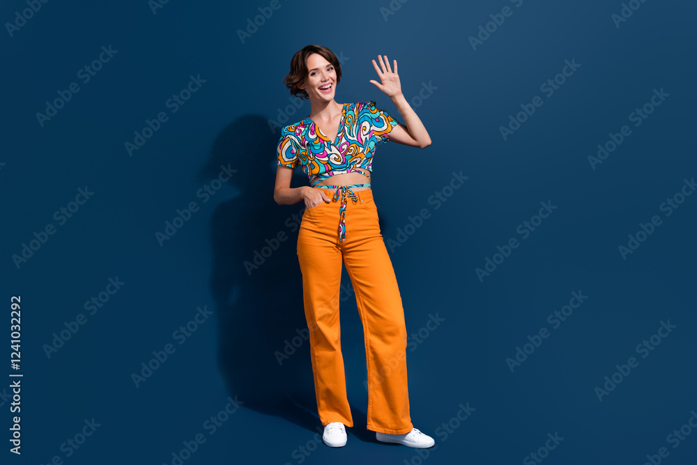 Fototapeta premium Full body photo of pretty young girl waving hand hello greeting dressed stylish flared pants outfit isolated on dark blue color background