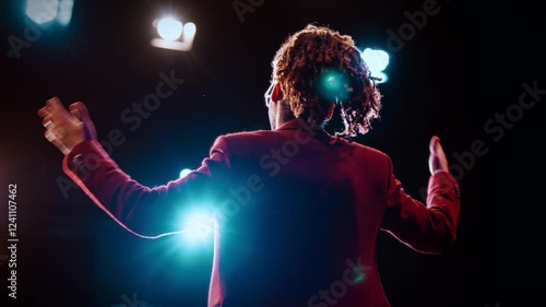 Speaker or host on stage of concert hall, back view against bright soffits. Rearview of charismatic african man with microphone standing alone in front of audience and speaking, motivating life coach